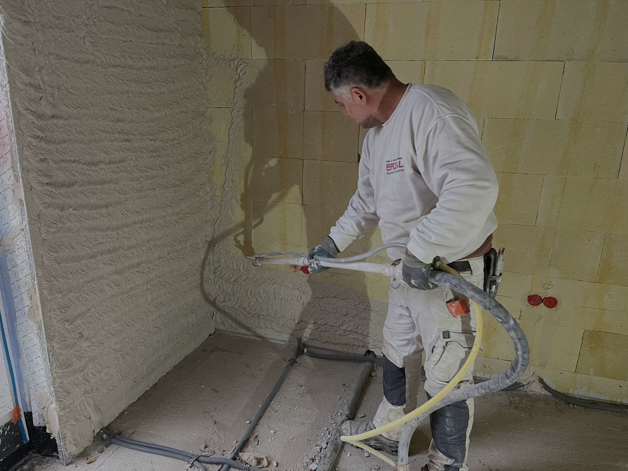 Ein Handwerker in Arbeitskleidung mit Erdal-Logo trägt Maschinenputz auf eine Innenwand auf. Links ist die Wand schon verputzt, rechts sind die Steine noch roh.